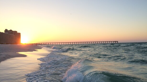 Ein Strand mit einem Steg, der sich ins Meer erstreckt, ein Hotel im Hintergrund und brechende Wellen am Ufer.