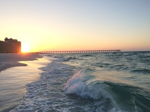 A beach with a pier extending into the ocean, a hotel in the background, and waves crashing onto the shore.