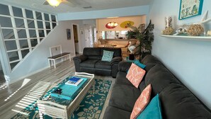 Ein Wohnzimmer mit einem dunkelbraunen Sofa, einem Couchtisch mit Tablett und einem gemusterten Teppich. Eine Treppe mit weißen Geländern führt zu einer weiteren Ebene.