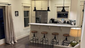 A modern kitchen with a refrigerator, bar stools, pendant lights, and a dining area with a table and chairs.