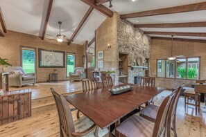 A spacious dining area with a large wooden table and chairs, a stone fireplace, and a view of the outdoors.
