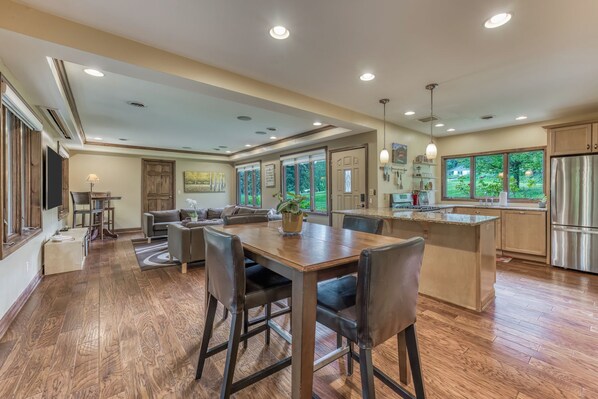A spacious kitchen with a dining area, wooden flooring, and large windows offering a view of the outdoors.
