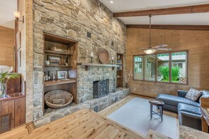 A cozy living room with a stone fireplace, wooden shelves, a blue leather sofa, and a large window with a view of greenery.