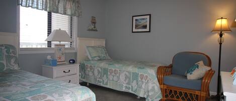 Un dormitorio con dos camas individuales, una silla, una mesita de noche, una lámpara y una ventana con persianas.