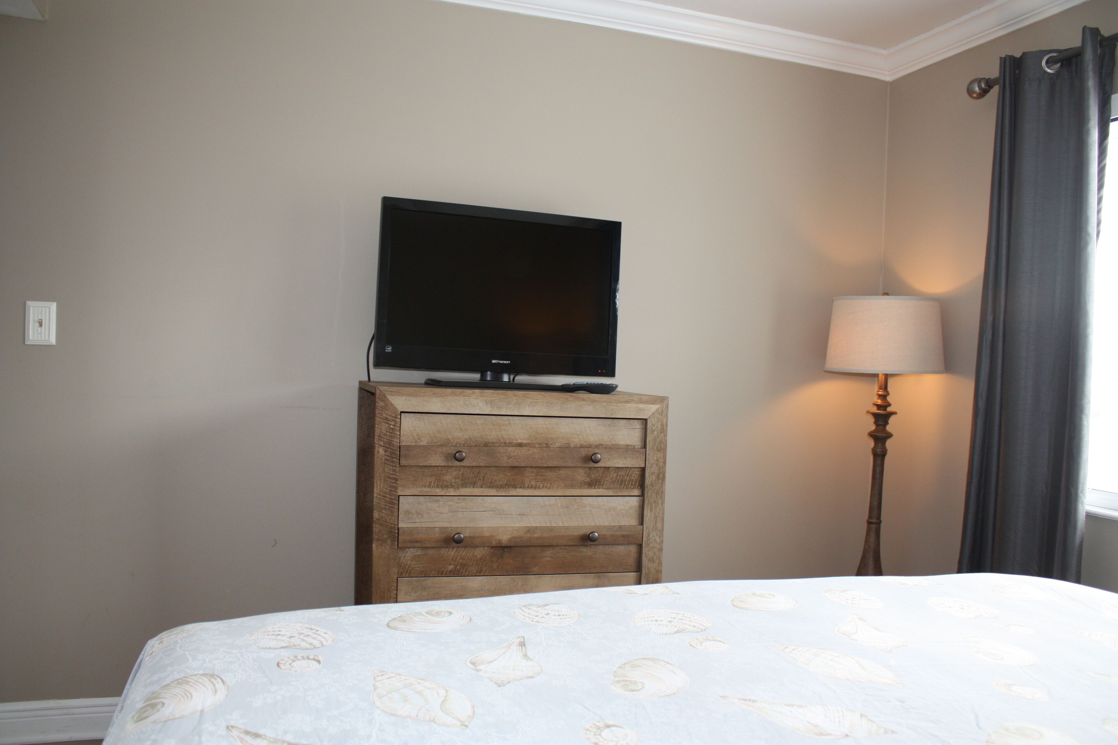 A bedroom with a bed, a wooden dresser, a television on top, and a lamp beside the window.