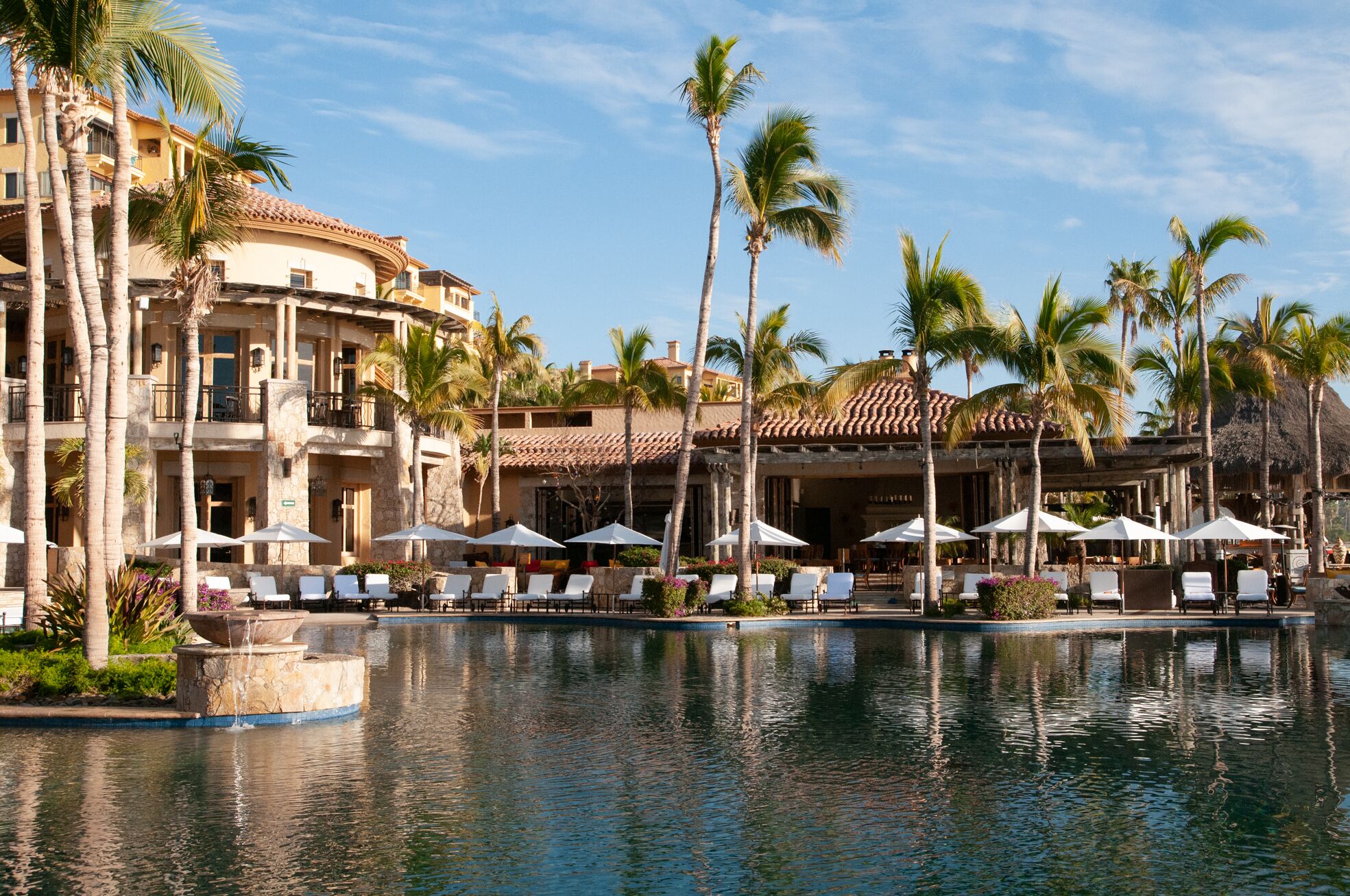 A resort with a pool, lounge chairs, umbrellas, and palm trees.