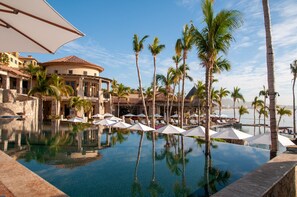 A poolside area with umbrellas, palm trees, and a building with a tiled roof. - Corner Unit, Building 4. Beautifully Remodelled (Cabo San Lucas)