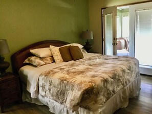 Une chambre à coucher avec un grand lit, une table de chevet, une lampe et une vue sur l’extérieur par une porte.