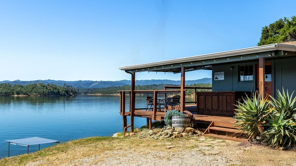 A lakeside cabin with a deck, outdoor seating, and a view of the lake and mountains.