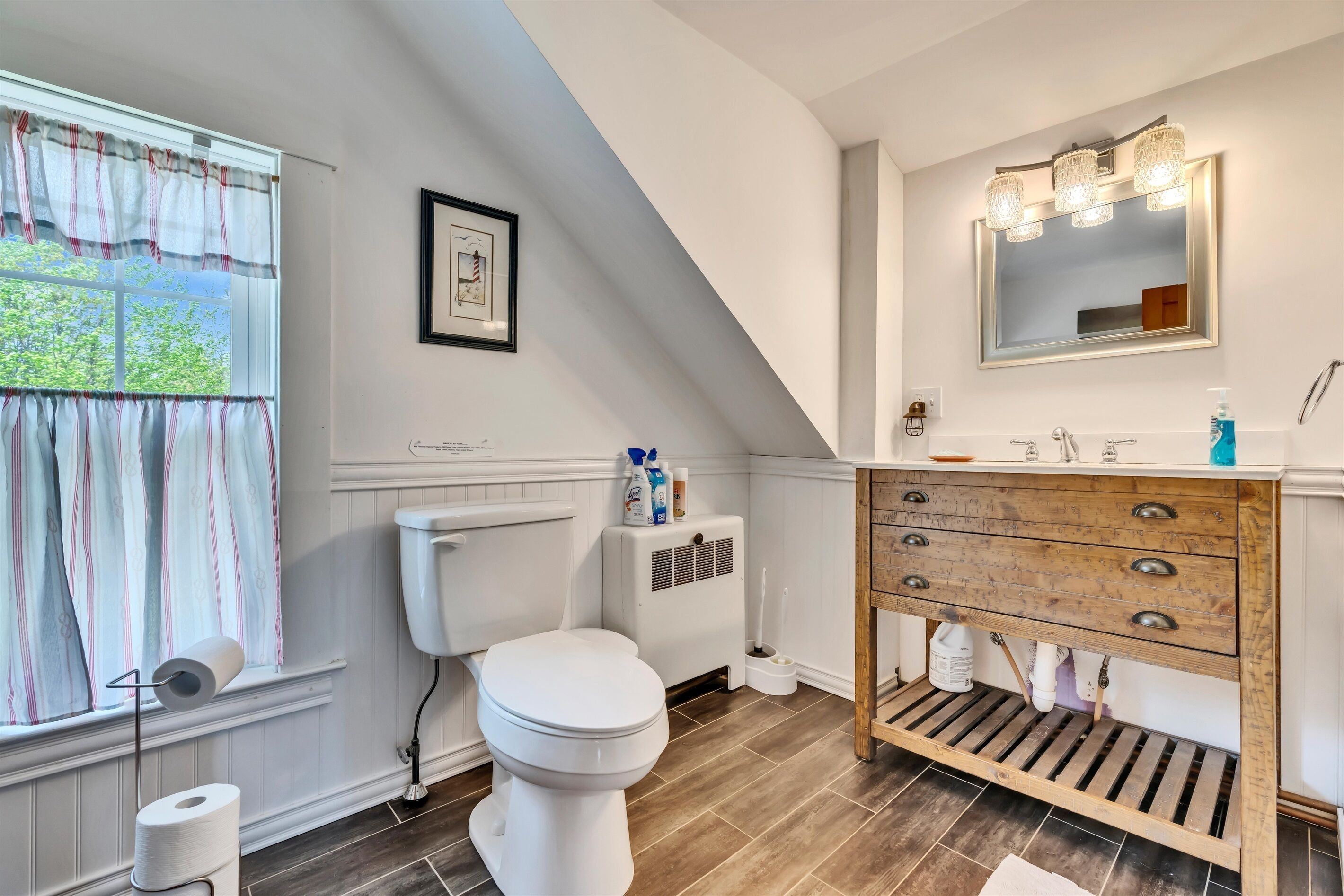 A bathroom with a toilet, a window with curtains, a sink with a mirror, and a wooden vanity.