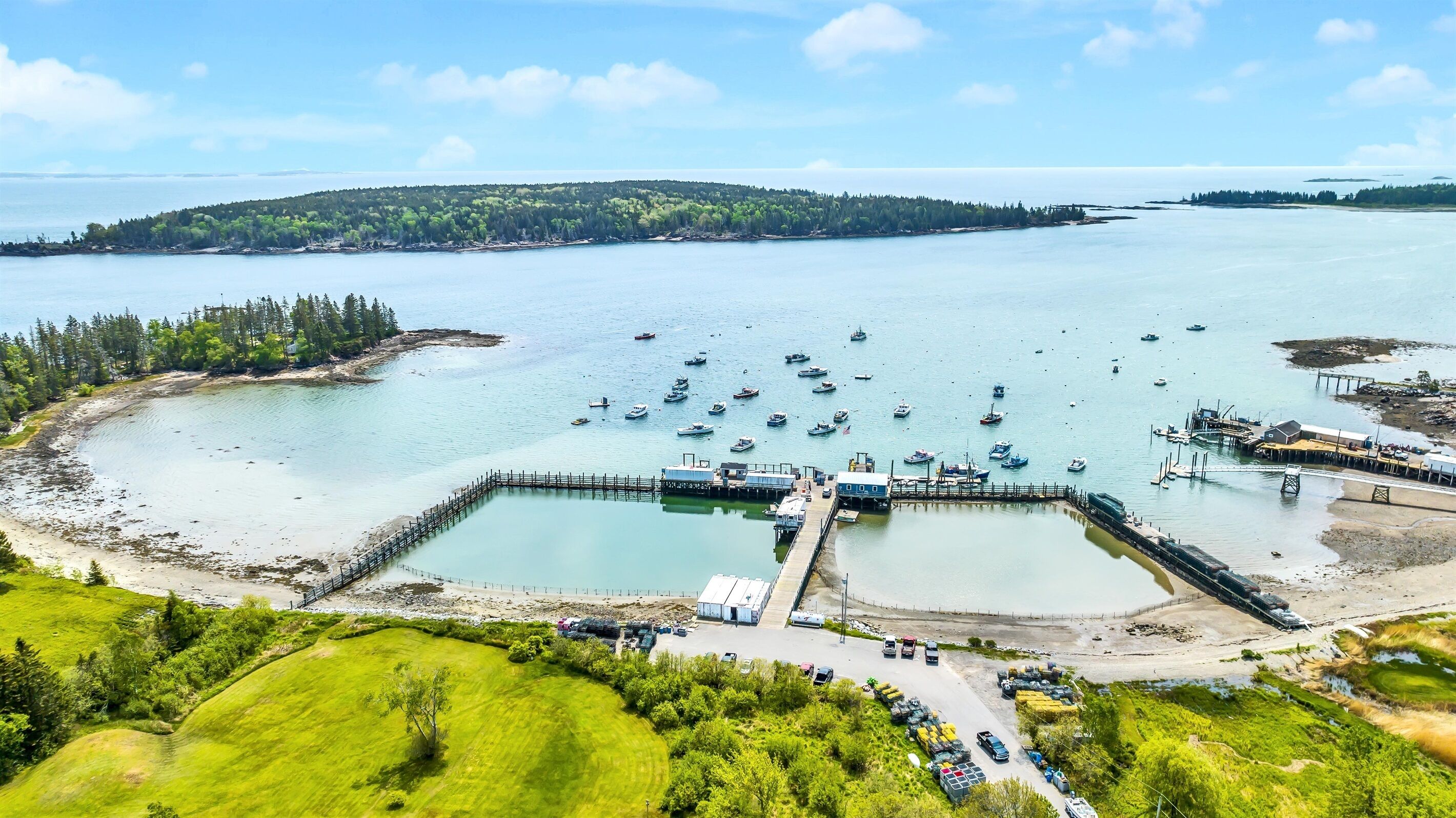 A coastal marina with multiple docks and boats, surrounded by green landscapes and a distant forested island.