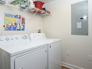 A laundry room with a front-loading washer and dryer, a shelf with various items, and a wall-mounted electrical panel.