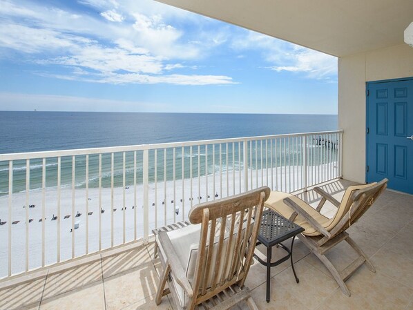 A balcony with a view of the beach and ocean. There are two wooden chairs and a small table.