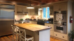 Una cucina con mobili in legno, elettrodomestici in acciaio inossidabile, un'isola in legno con sedie, luci a sospensione e una finestra che lascia entrare la luce del giorno.