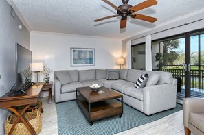 A living room with a grey sectional sofa, a wooden coffee table, a TV, and a ceiling fan. - Firethorn 822- 2 bedrooms 1.5 baths overlooking canal w/private beach access (Siesta Key)
