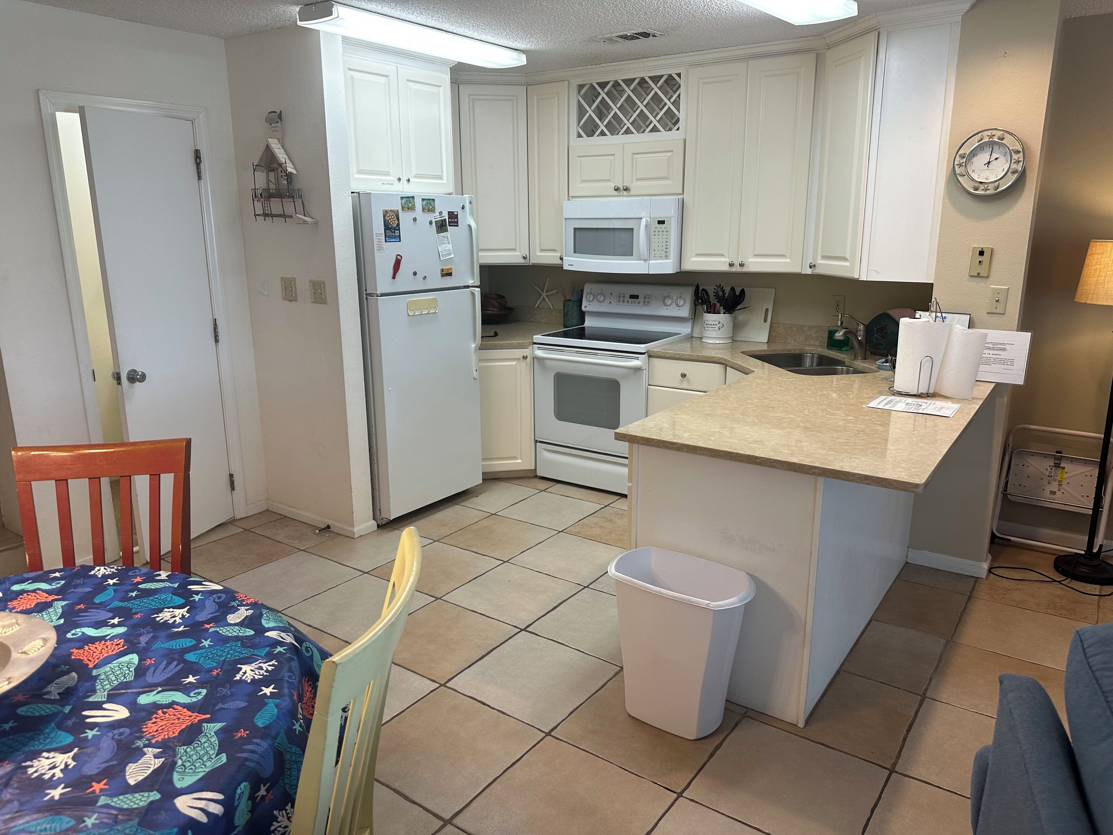 A compact kitchen with white cabinets, a refrigerator, a microwave, an oven, and a sink. There is a dining table with a colorful tablecloth and a chair. A trash bin is placed near the sink.