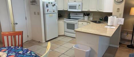 A compact kitchen with white cabinets, a refrigerator, a microwave, an oven, and a sink. There is a dining table with a colorful tablecloth and a chair. A trash bin is placed near the sink.