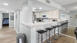 Une cuisine moderne avec des armoires blanches, un îlot central doté de tabourets de bar, des appareils en acier inoxydable et une vue sur la chambre adjacente.