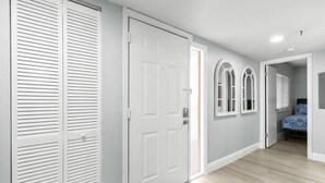 Un couloir avec une porte blanche, un placard avec des portes à persiennes, et une pièce avec un lit et une literie à motifs.