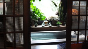 Une vue à travers une fenêtre ornée d’un treillage en bois, avec un élément décoratif d’eau, des plantes en pot et un vase décoratif.