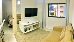 A living room with a TV on a white stand, a beige sofa, a patterned bed, and a window with a city view. - Vacanze - edf. Gales de Pajuçara apt 204, rooftop pool and gym. (Ponta Verde)