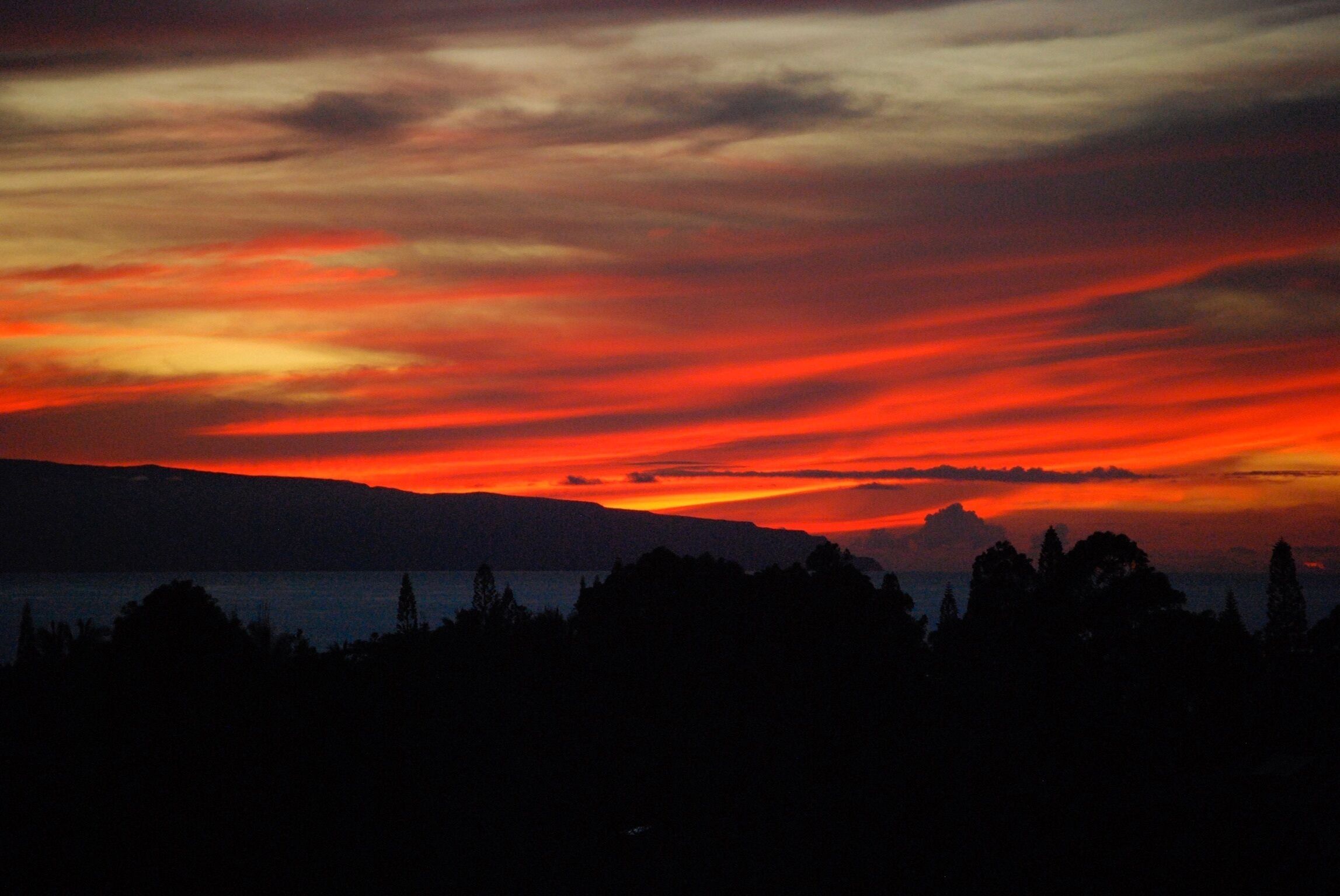 A sunset over a mountainous landscape with silhouettes of trees and a body of water.