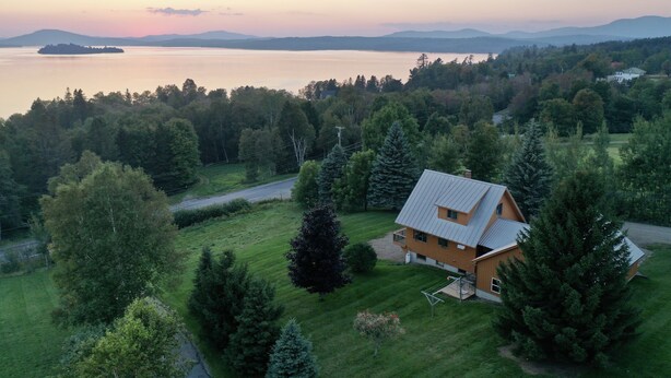 Une maison avec un toit en pente, entourée d’une pelouse bien entretenue et de nombreux arbres, qui offre une vue sur une vaste étendue d’eau et sur des montagnes lointaines.