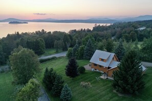 Une maison avec un toit en pente, entourée d’une pelouse bien entretenue et de nombreux arbres, qui offre une vue sur une vaste étendue d’eau et sur des montagnes lointaines.