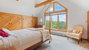 Une chambre à coucher avec une tête de lit en bois, une grande fenêtre donnant sur un beau paysage, un lit avec une literie blanche et un fauteuil.