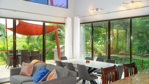 A modern dining area with a large table and chairs, a sofa with cushions, and large windows offering a view of lush greenery. - Private Rainforest Retreat with Infinity Pool & Concierge (Quepos)