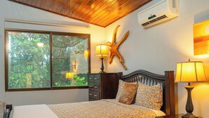 A bedroom with a bed, a nightstand, a dresser, a starfish wall decoration, and a window with a view of greenery. - Private Rainforest Retreat with Infinity Pool & Concierge (Quepos)