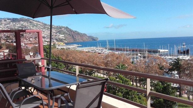 A balcony with a table and chairs, overlooking a marina with boats and a coastal town. - Superb View Over the Marina Funchal 270º view (Funchal)