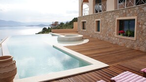 A pool area with a wooden deck, stone walls, and a view of the sea. - Breathtaking mediterranean view, large private infinite Pool & Jacuzzi ! (Theologos)