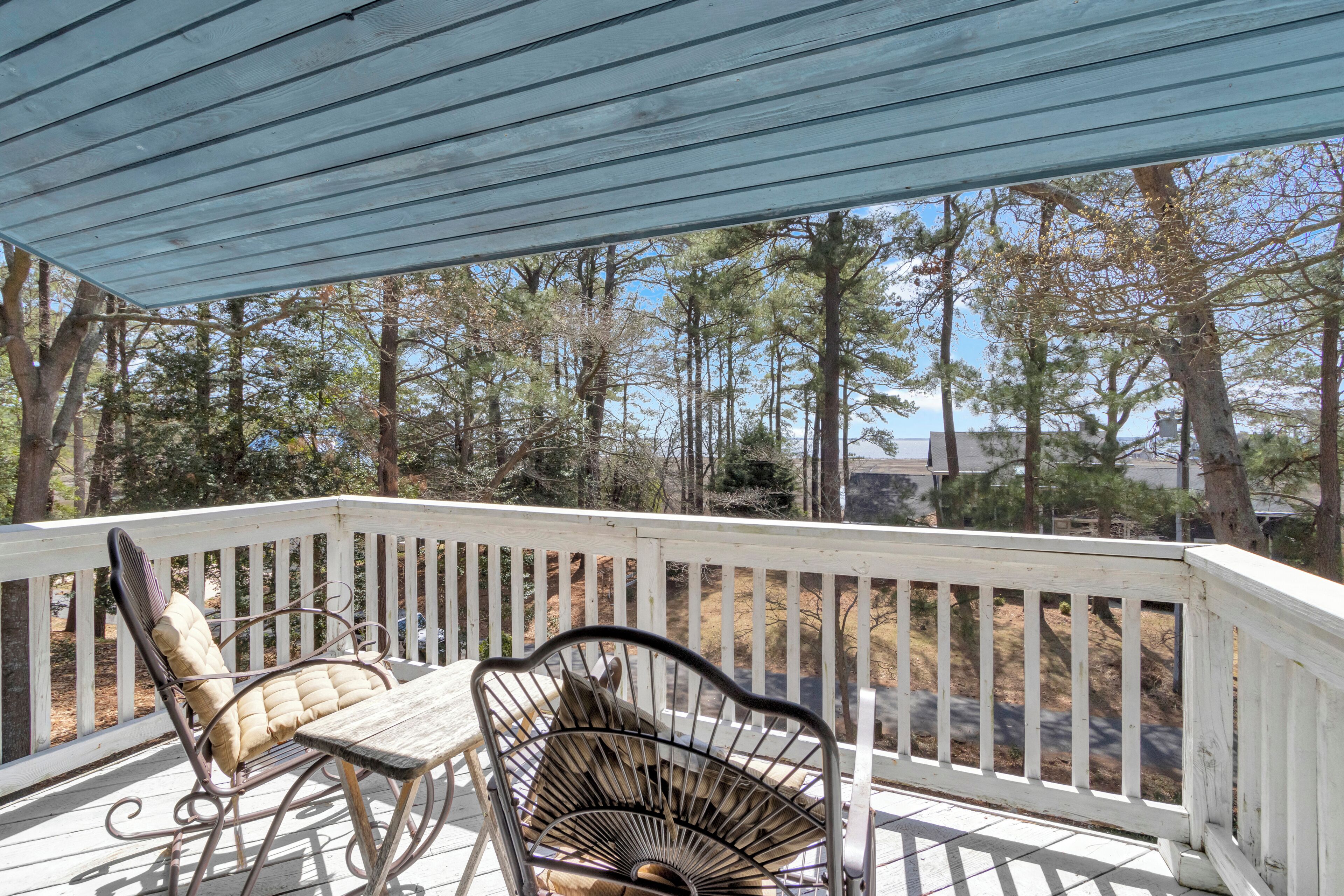 A covered outdoor deck with two metal chairs and a table, surrounded by a white railing, overlooking a forested area.