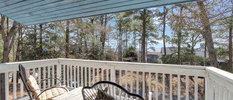A covered outdoor deck with two metal chairs and a table, surrounded by a white railing, overlooking a forested area.
