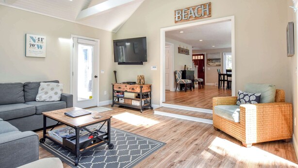 A spacious living area with a grey sofa, a wicker armchair, a coffee table, and a TV mounted on the wall. A sign reading "BEACH" is visible on the wall. - Amazing Backyard, Spacious, Dog Friendly! (Dennis)