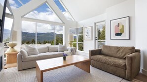 A modern living room with a high ceiling, large windows offering mountain views, a beige sofa, a brown armchair, and a wooden coffee table. - LUXURY- 2BA/BA True Ski-in/Ski-out, HotTub&Pool (Whistler)