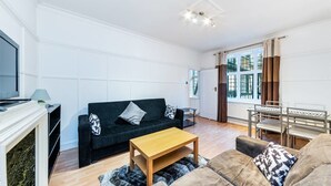 Ett modernt vardagsrum med en mörkgrön soffa, ett soffbord i trä och en beige soffa. Det finns en tv på ett tv-bord, ett litet sidobord och ett fönster med gardiner.