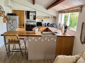 A compact kitchen with a wooden table, a chair, a refrigerator, a microwave, and a window with a view of greenery.
