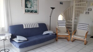 A living room with a blue sofa, striped pillows, a wooden chair, a floor lamp, and a staircase. - Apartment in a small quiet residence near the sea (Château-D'olonne)