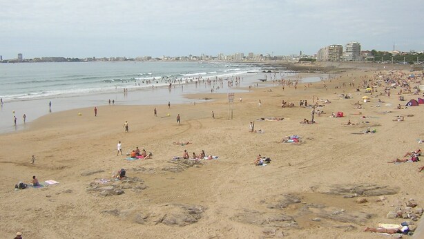 A wide sandy beach with people sunbathing and swimming, a clear sky, and buildings in the distance. - Apartment in a small quiet residence near the sea (Château-D'olonne)