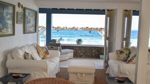 Un coin salon accueillant avec deux canapés blancs et un petit pouf, une chaise en osier et une table sur laquelle se trouve un saladier. On peut voir un mur de pierre et une vue sur l’océan, avec des personnes sur la plage.