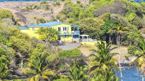 Une maison jaune avec un toit bleu, entourée d’une végétation luxuriante et de palmiers, et qui donne sur une étendue d’eau.