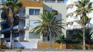 A multi-story building with balconies, palm trees, and a sandy ground.