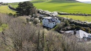 A white two-story house with a sloped roof, surrounded by a dense forest and a green field. - Schoopers is set amongst rolling green fields on the edge of the Camel Estuary. (Wadebridge)