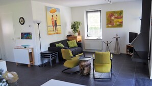A modern living room with a sofa, chairs, a coffee table, a small side table, a lamp, a plant, a painting, and a television.