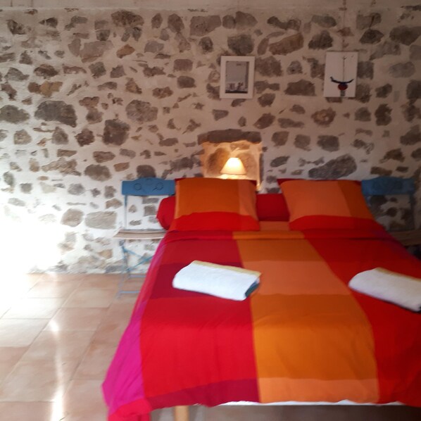 A bed with a red and orange bedspread, a lamp on the wall, and a stone wall. - Large Provencal country house in a wooded park with very large swimming pool (Lorgues)