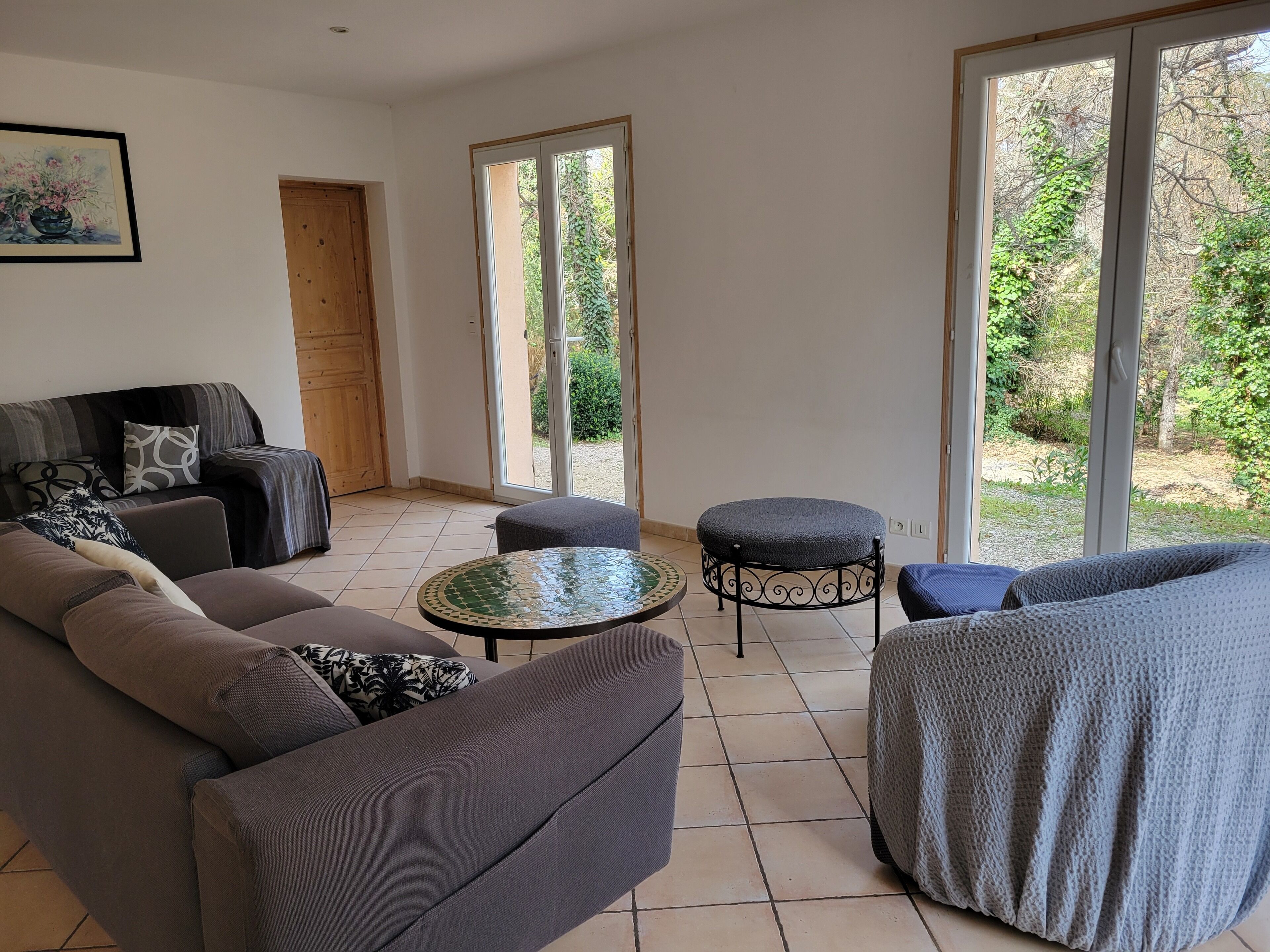 A living room with a sofa, armchairs, a small round table, and a view of greenery through large windows.