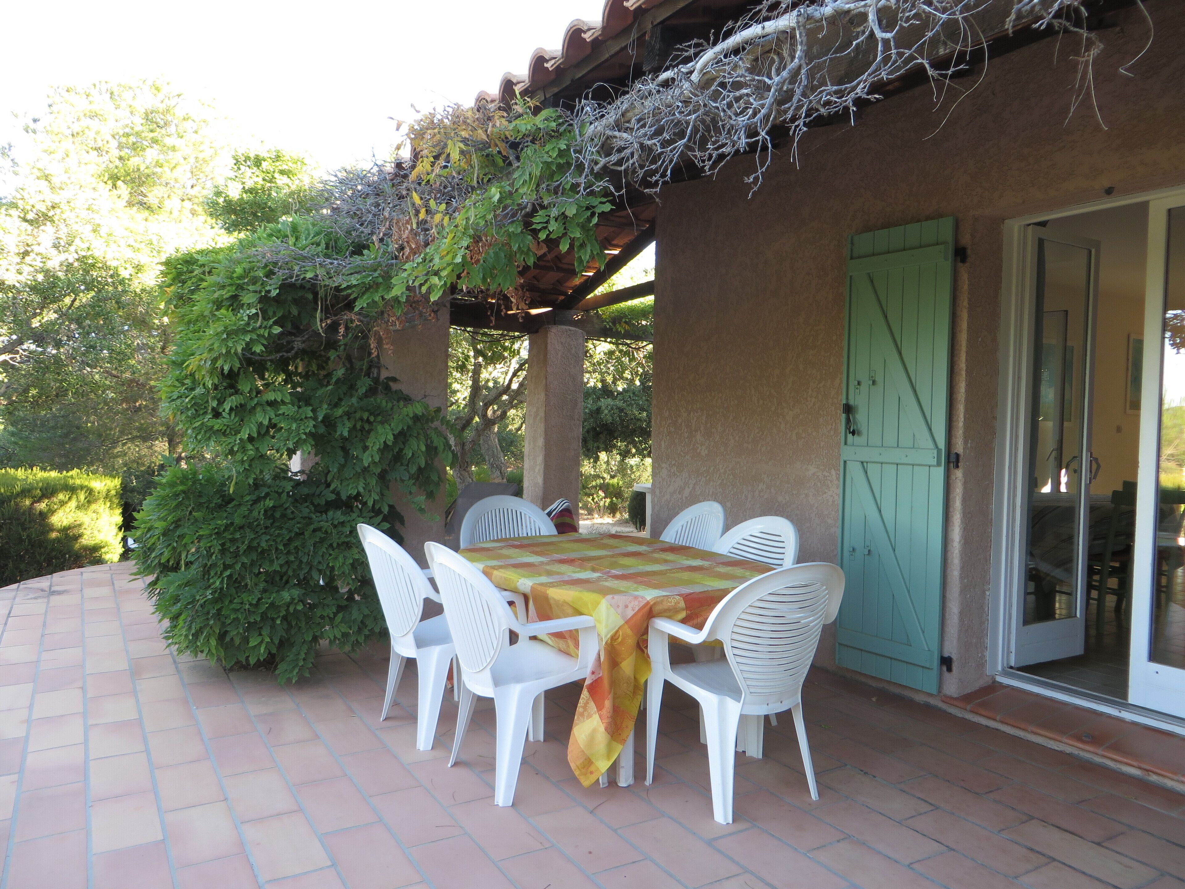 Une terrasse extérieure avec une table et des chaises, un espace abrité avec un toit, et une porte donnant sur une entrée couverte.