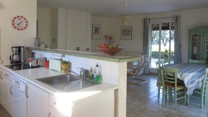 Een keuken met een spoelbak, een koffiezetapparaat en een fruitschaal op het aanrecht. Er is een eettafel met stoelen en een raam met gordijnen.